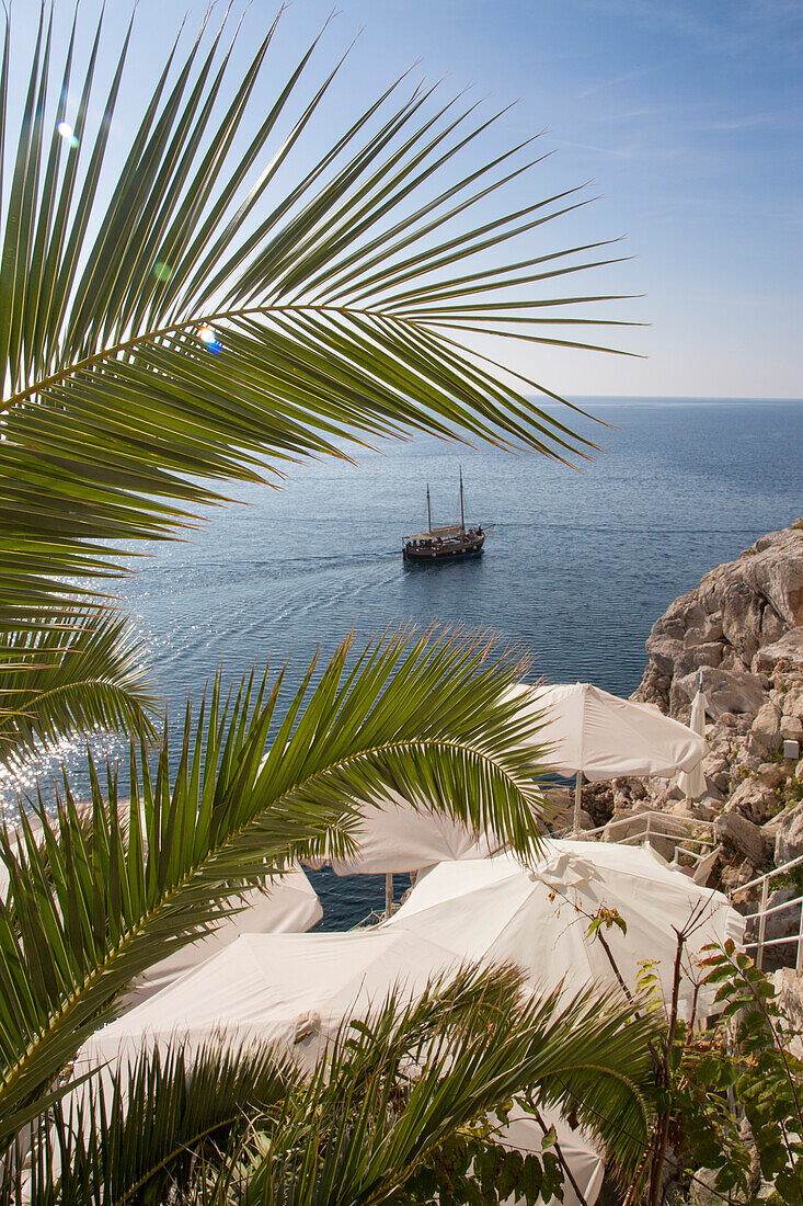 Palmwedel, Sonnenschirme einer Bar und Ausflugsboot nahe Küste, Dubrovnik, Dalmatien, Kroatien, Europa