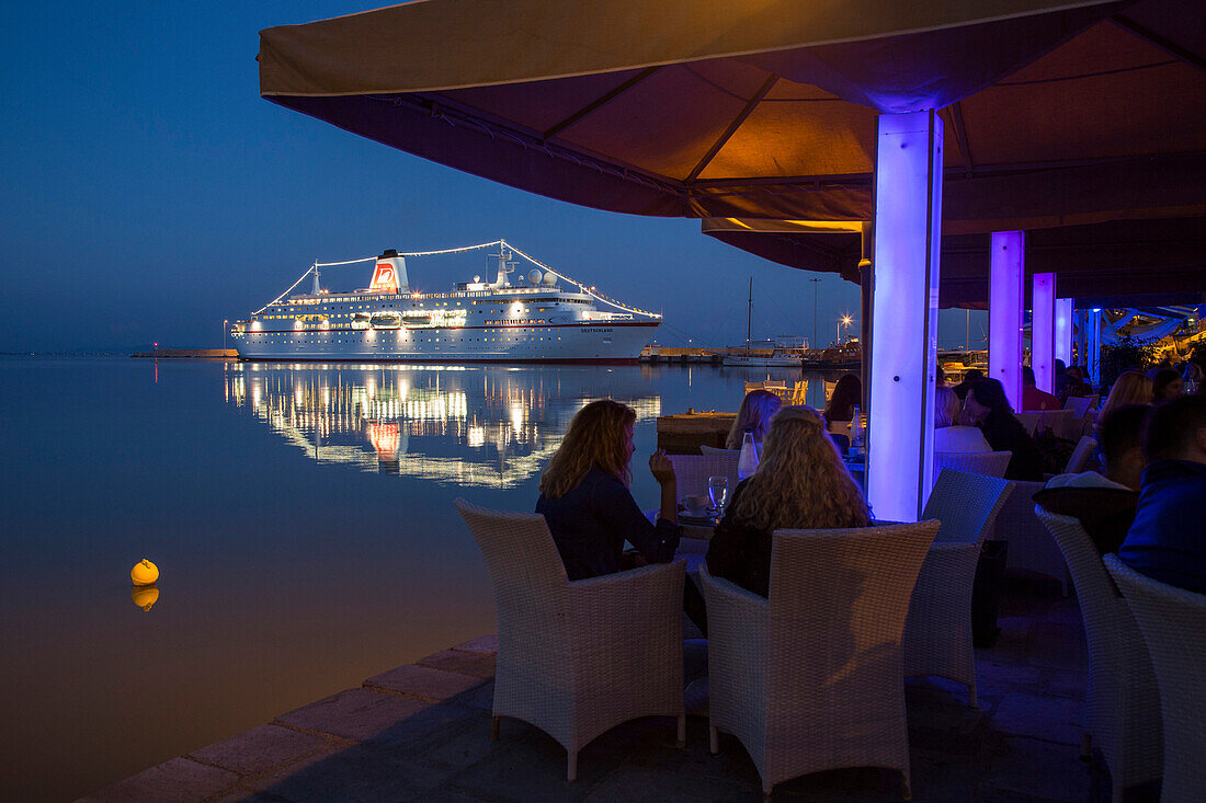 Menschen sitzen in Restaurant mit Blick auf Kreuzfahrtschiff MS Deutschland, Reederei Peter Deilmann, an der Pier im Dämmerlicht, Katakolon, Pyrgos, Peloponnes, Griechenland, Europa