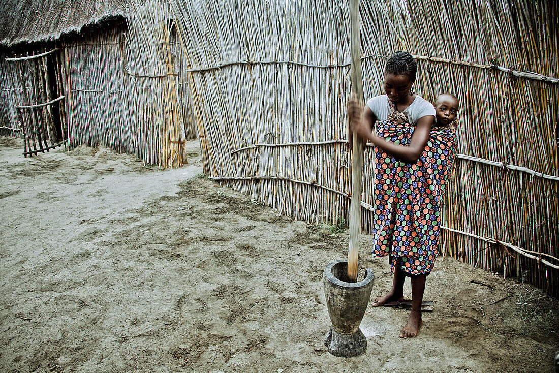 Young mother from the Lozi tribe with baby pounding grain in a big wooden mortar, Caprivi region, Namibia, Africa