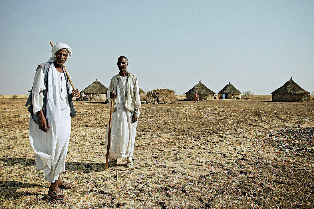 Two men in jellabiya in front of a village with round huts, East Sudan, Africa
