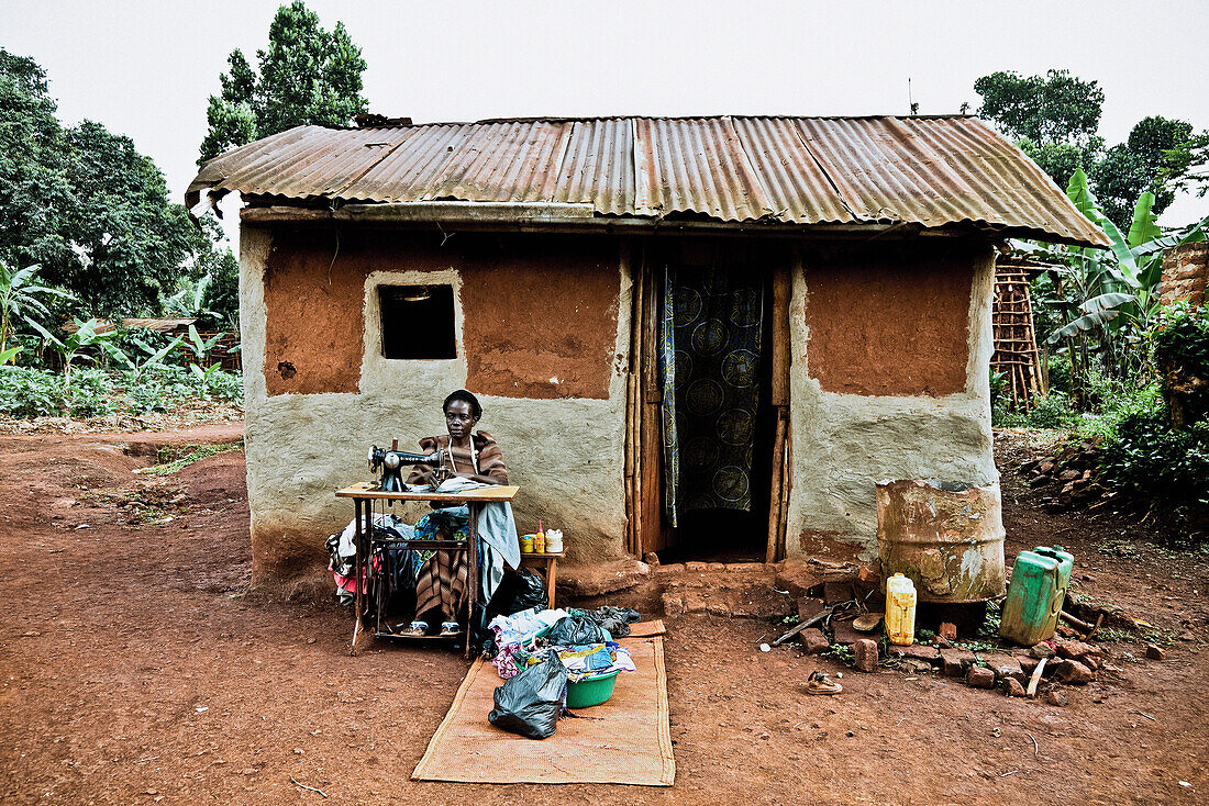 Frau sitzt vor ihrer Hütte an einer Nähmaschine, Buwenda, Uganda, Afrika