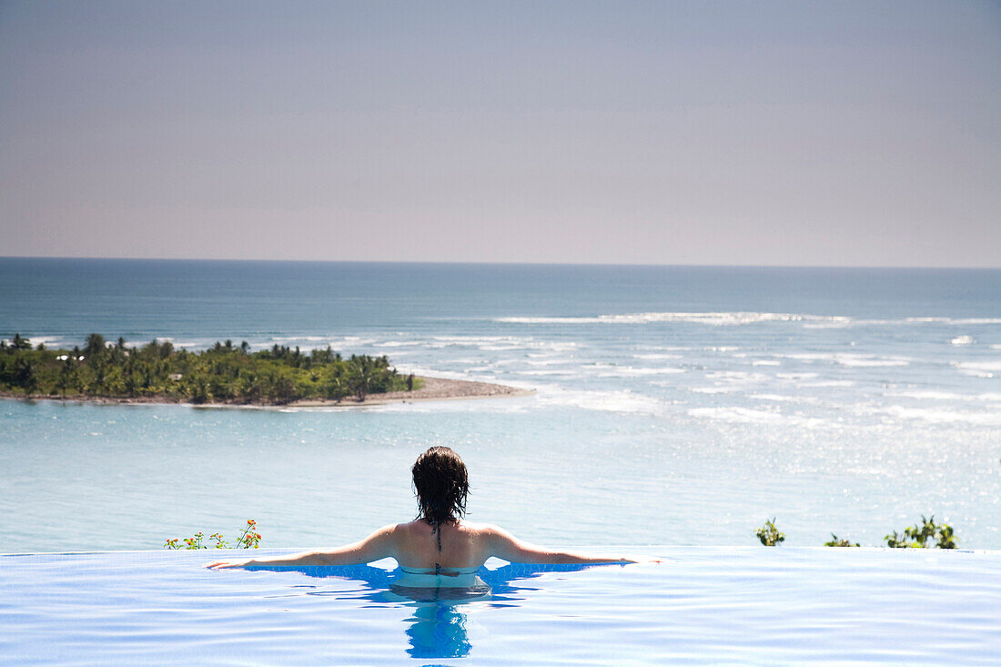 A woman stands in an infinity pool with arms stretched over the edge looking at the blue ocean beneath Ojochal, Costa Rica