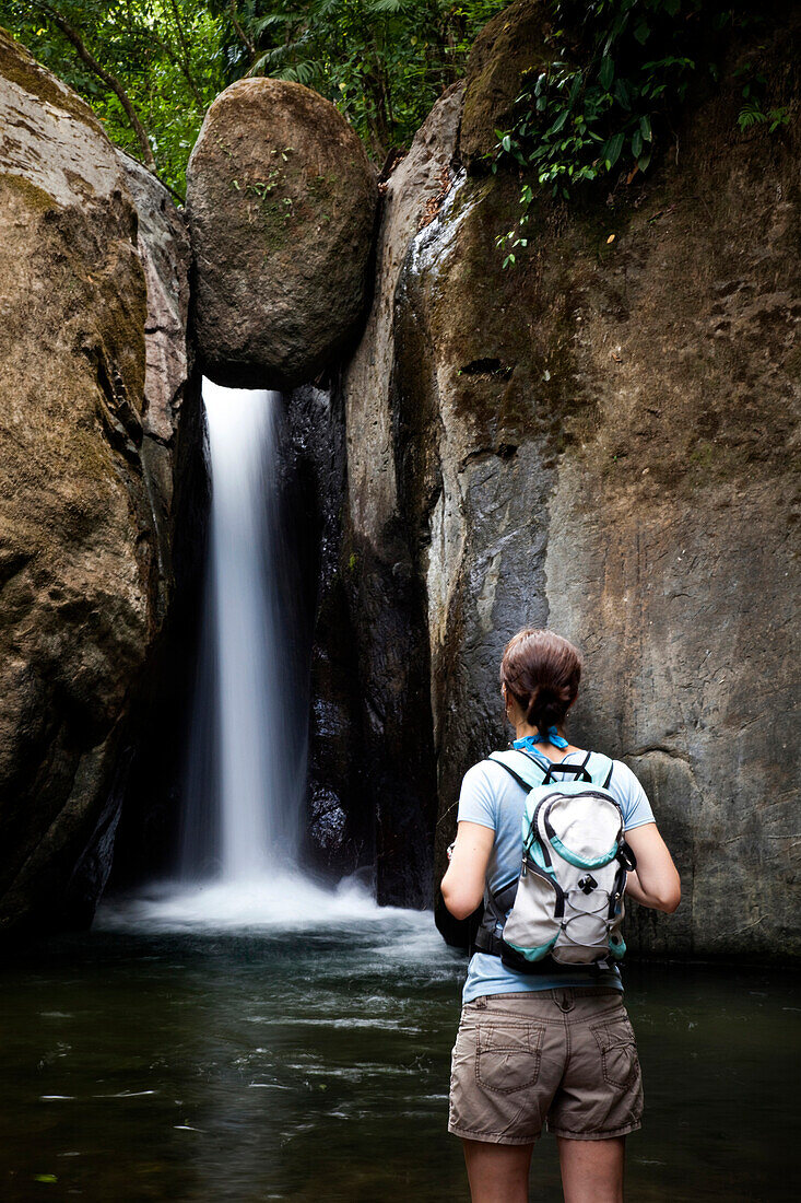 A woman stands at the edge of a pool of water looking at a waterfall while wearing a backpack Costa Rica