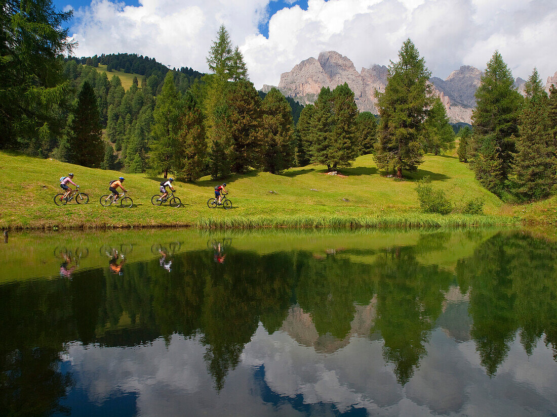 Four mountain bikers are riding a grassy slope along a mountain lake with rock cliffs in the background Val Gardena, Dolomites, Italy