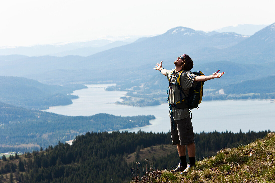 A young man holds his arms open in success after hiking to the top of a mountain in Idaho Sandpoint, Idaho, USA