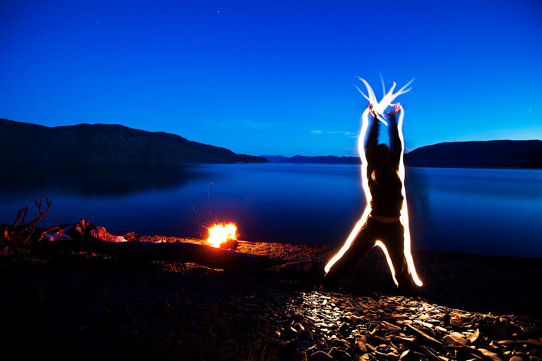 A woman sends energy through her body and out her hands at sunset next to a campfire and lake in Idaho. This light painting image was created with a long exposure and flash lights Sandpoint, Idaho, USA