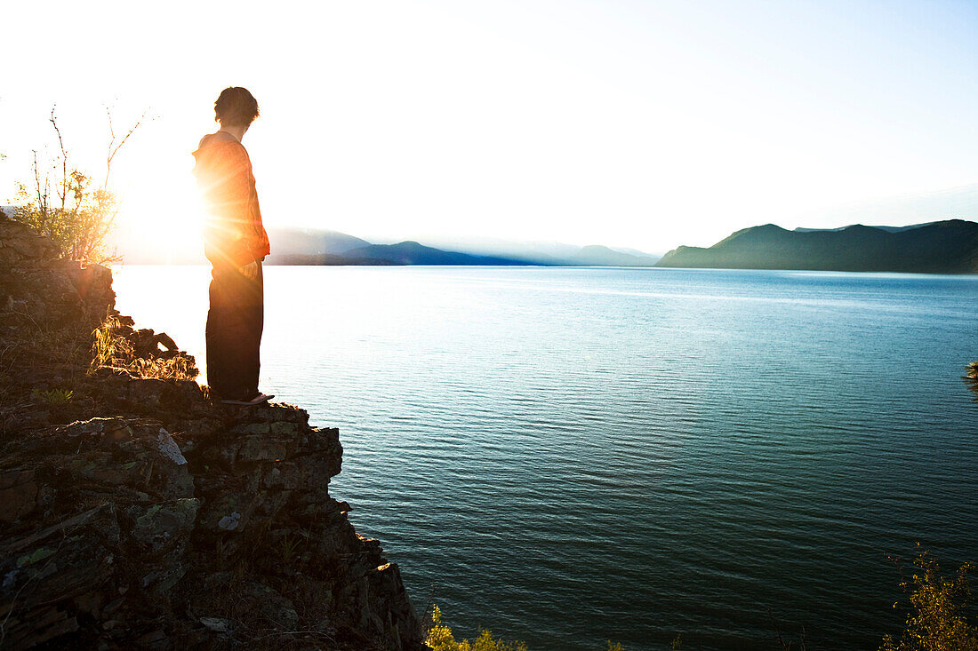 A handsome man peacefully stands on the edge of a cliff watching the sun rise over the lake in Idaho Sandpoint, Idaho, USA