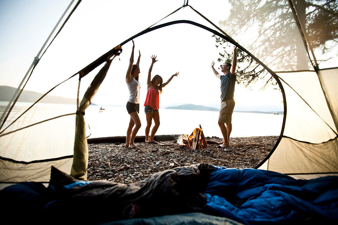 Three young adults smiling and dancing around a camp fire on a camping and kayaking trip on a lake in Idaho Sandpoint, Idaho, USA