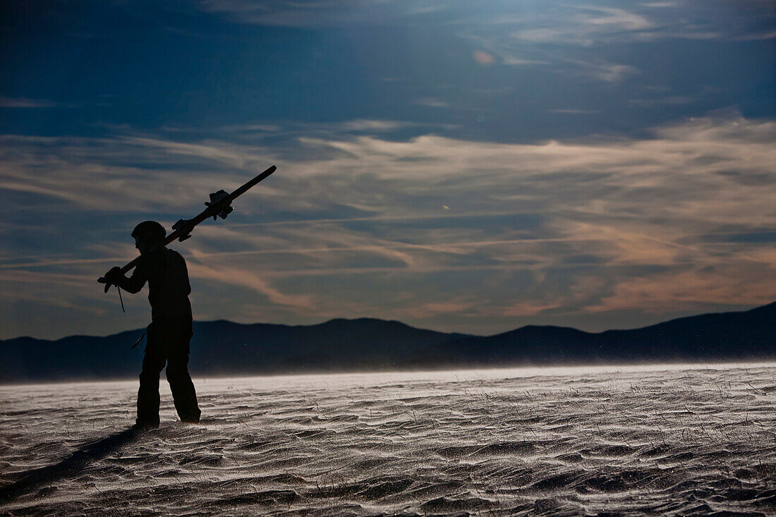 A man stands with his skis on a snow covered mountain silhouetted by the sun., Madison County, NC, United States
