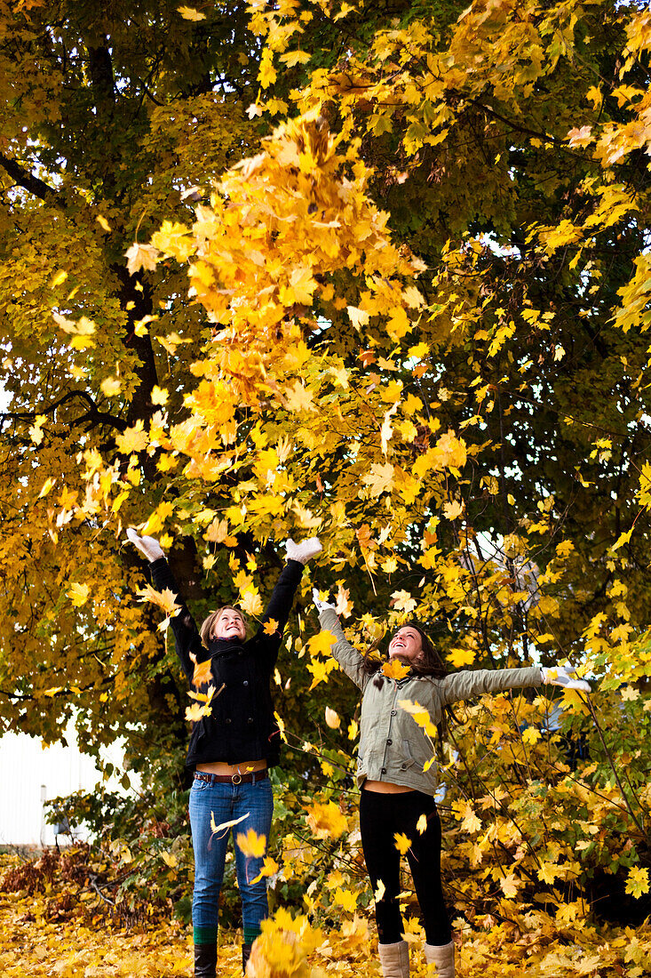 Two young women smiling throw orange leaves into the sky surrounded by fall colors in Idaho., Sandpoint, Idaho, USA