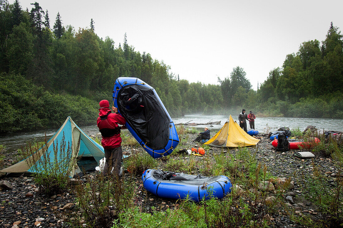 Four friends packing up camp by a river with tents and packrafts in the rain., Talkeetna, AK, USA