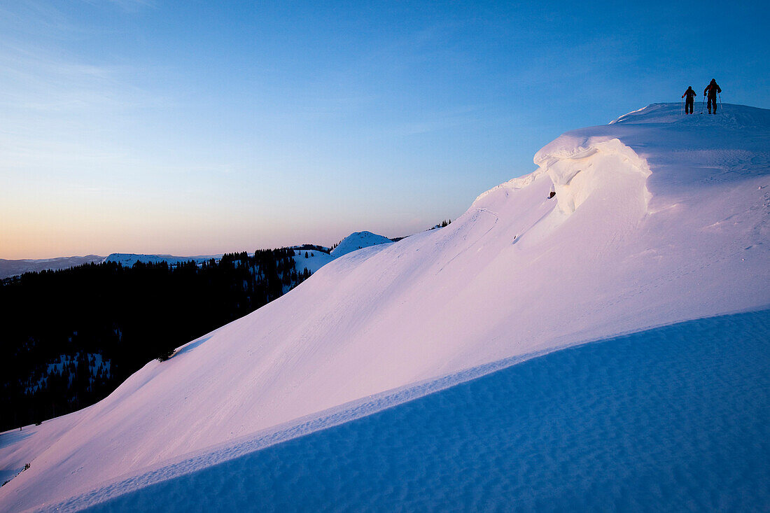 Silhouettes of two people backcountry skiing up a mountain at sunrise in purple light., Park City, Utah, USA