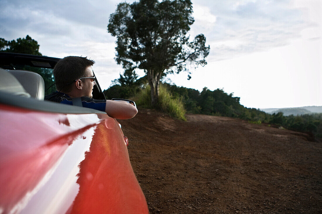 A young man looks out from the passenger seat of a red convertible sports car in Hawaii., Waimea, HI, USA