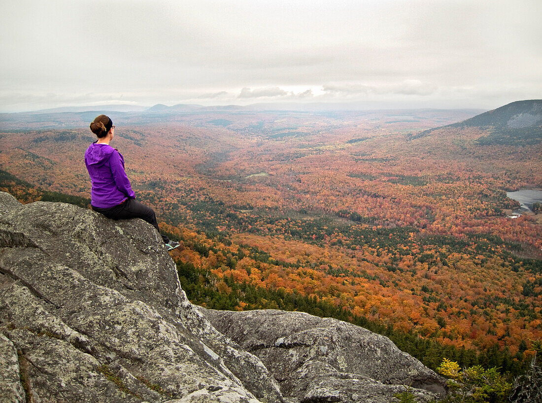 A young female hiker views vibrant fall foliage from the rocky summit of a New England peak., Elliotsville Plt., ME, USA