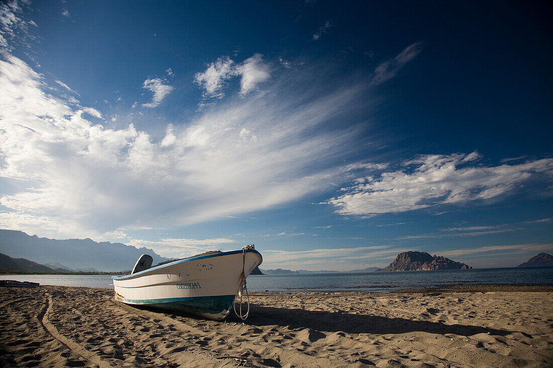 A boat sits on the sand on an empty beach beneath a partly cloudy sky in Loreto, Baja California Sur, Mexico Loreto, Baja, Mexico