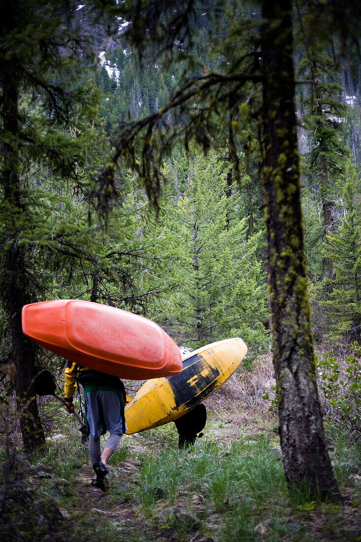 Two kayakers carry their boats through a dark forest to the put-in Mazama, Washington, USA