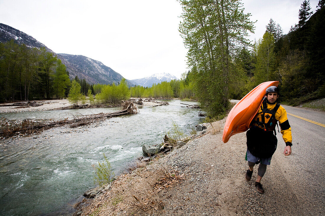 A young man carries a kayak down a road with a river alongside him Mazama, Washington, USA