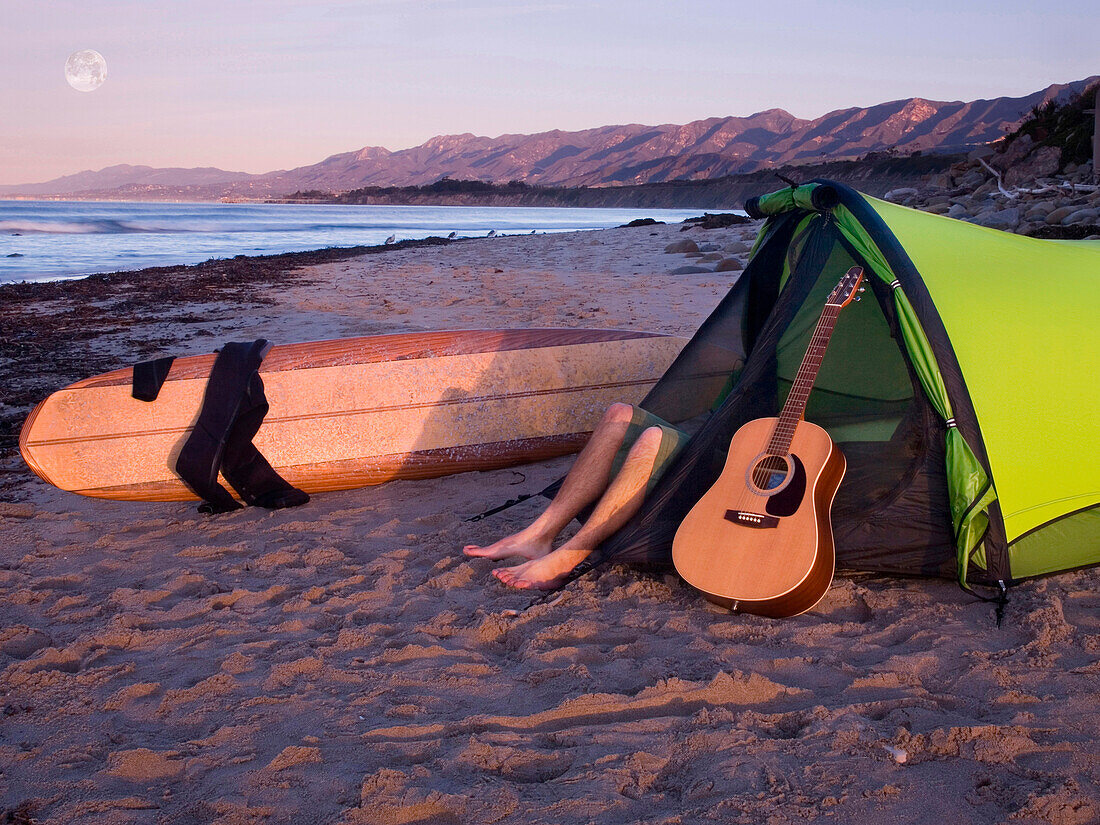 A young man lying in his tent with a guitar while camping on the beach waiting for surf Carpinteria, California, USA