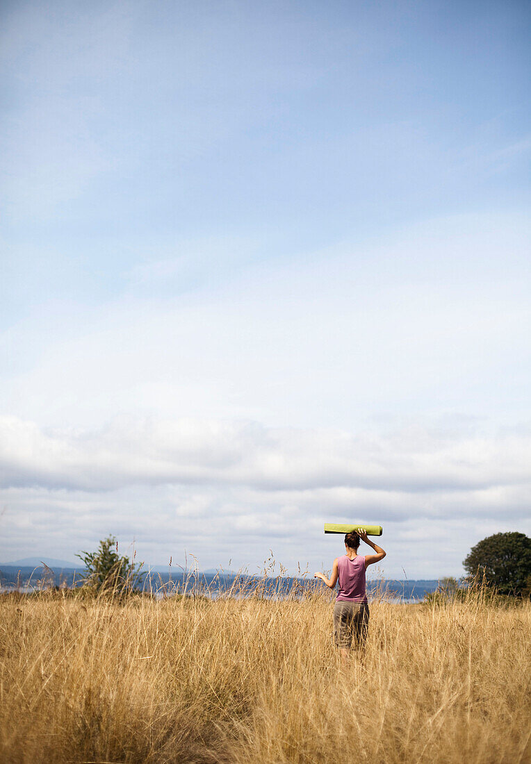 A young woman, wearing a purple shirt, carries a green yoga mat through a field of golden grasses overlooking the ocean Seattle, Washington, USA