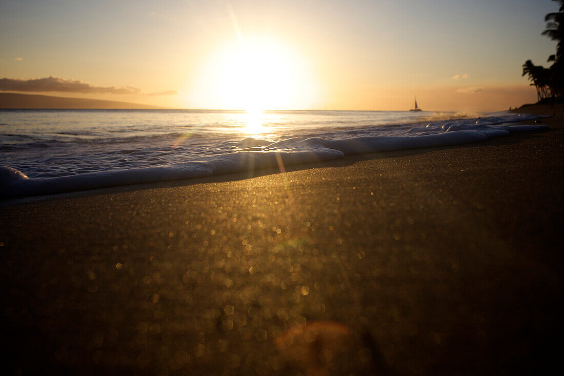 Low angle view of a beach at sunset highlights the foam of a wave coming into the sand as a boat passes by in the background Maui, Hawaii, USA
