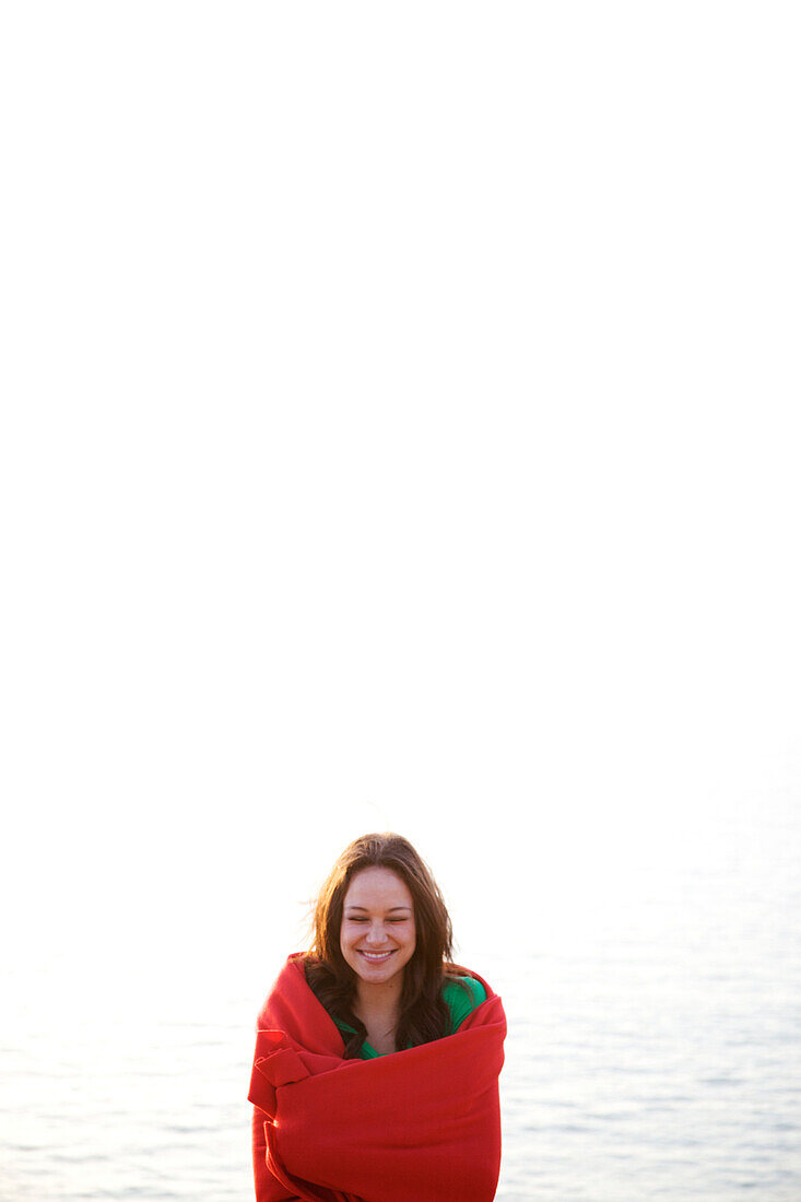 Female in her early 20's wrapped in a red blanket smiles with her eyes closed on the beach San Diego, California, USA