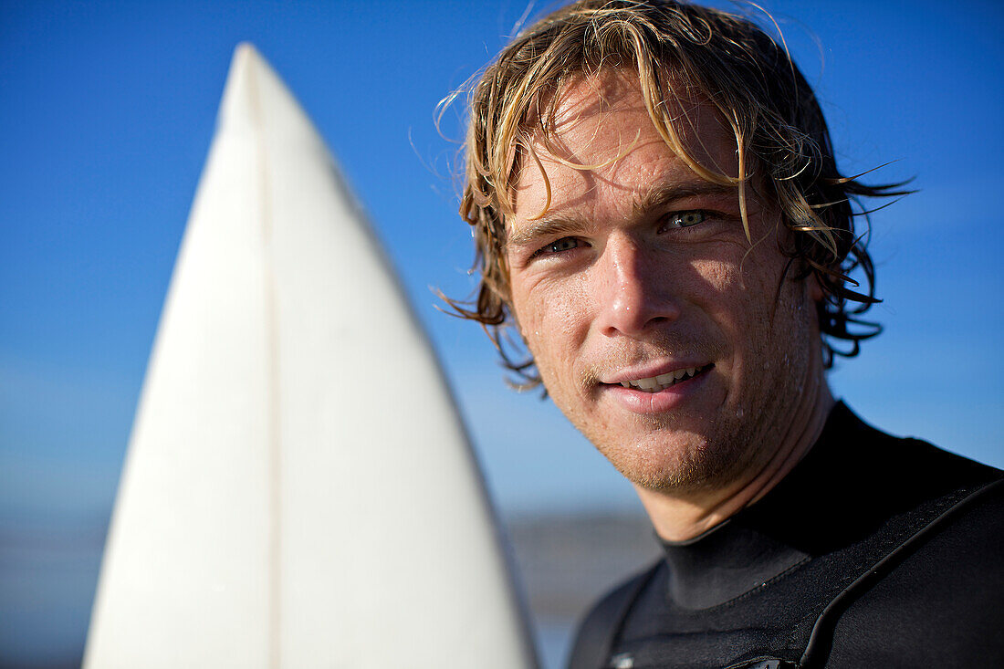 Young male surfer with wet hair at Pacific Beach in San Diego, California on a sunny day looks straight at the camera with his surfboard behind him San Diego, California, USA