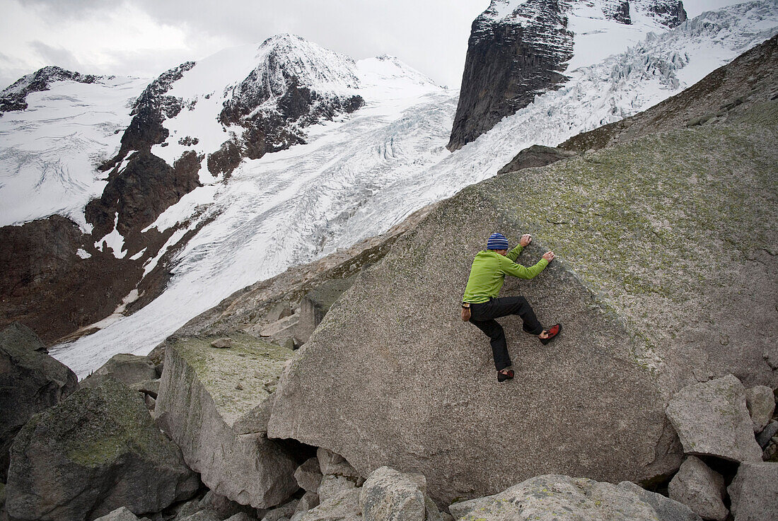 Man wearing a green shirt climbs a granite boulder with large glacier in background British Columbia, Canada