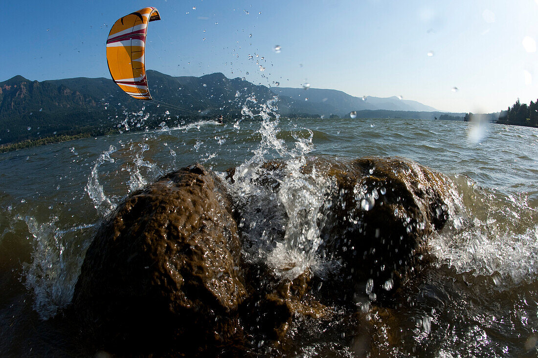 Low angle perspective of a kiteboarder riding by with waves splashing on a rock prominent in the foreground Stevenson, Washington, USA