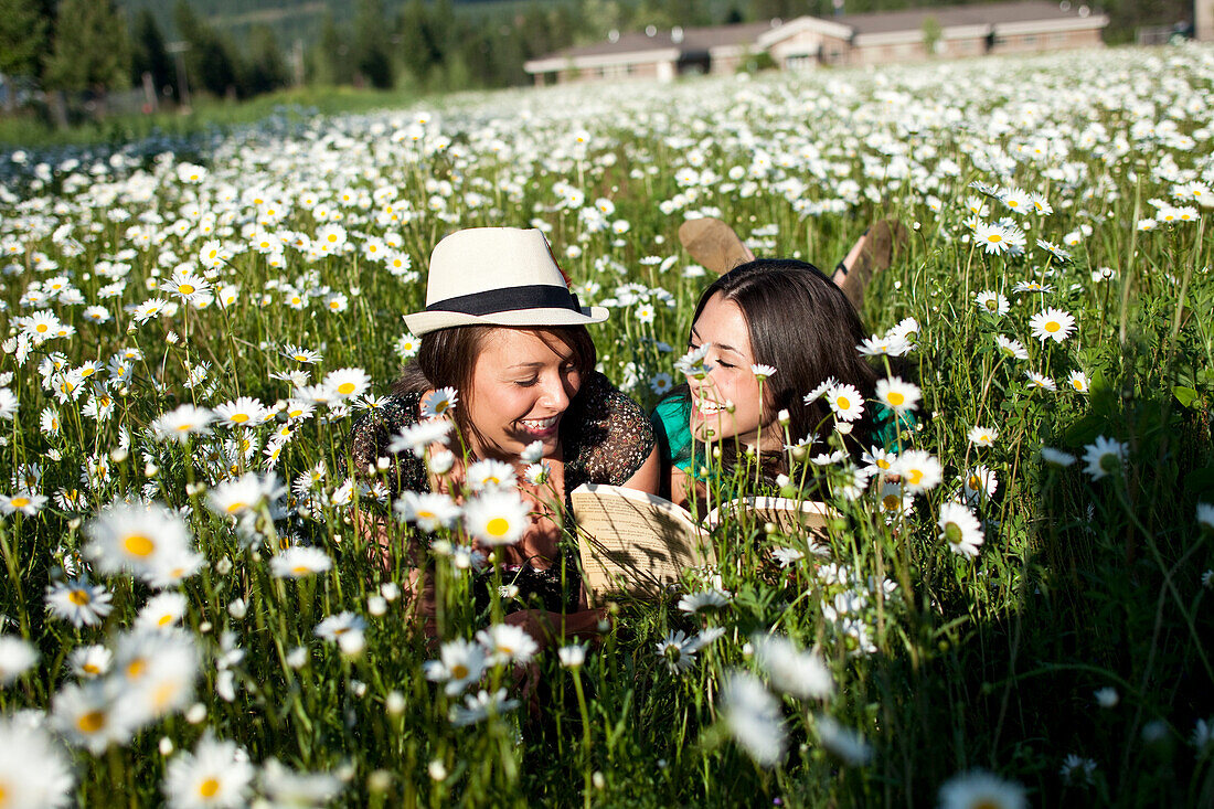 Two young women smile while reading a book in a field of wild flowers Sandpoint, Idaho, USA