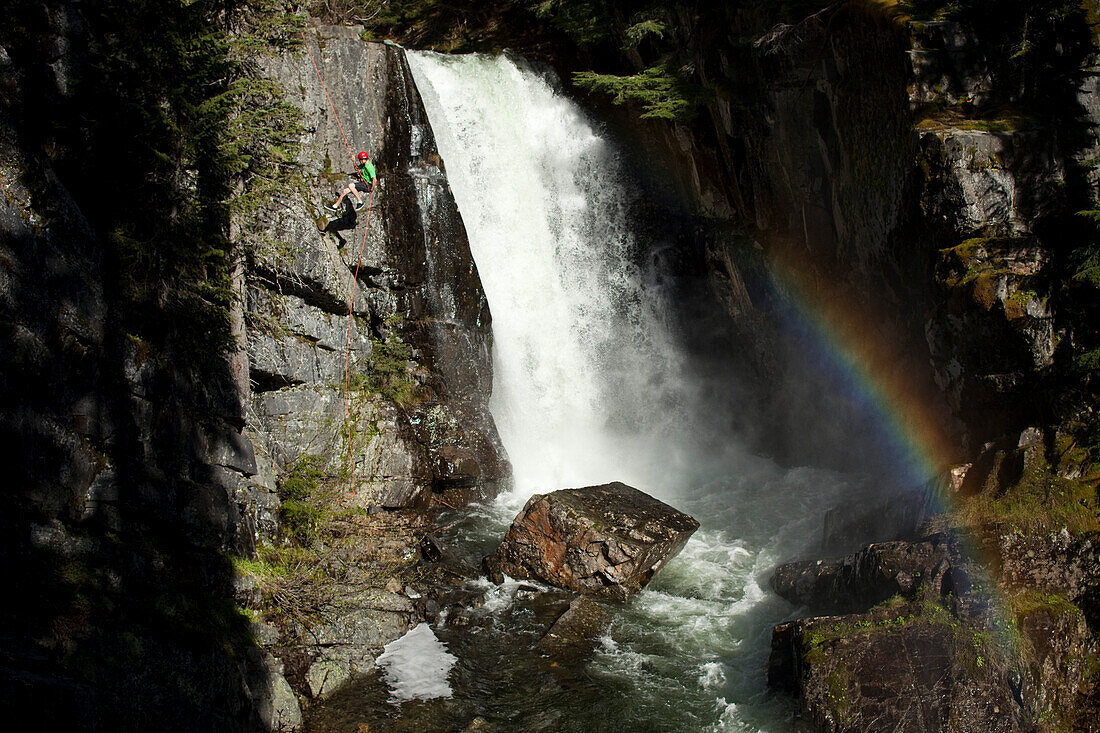 A young man rappels down a cliff next to a waterfall and rainbow in Idaho Sandpoint, Idaho, USA