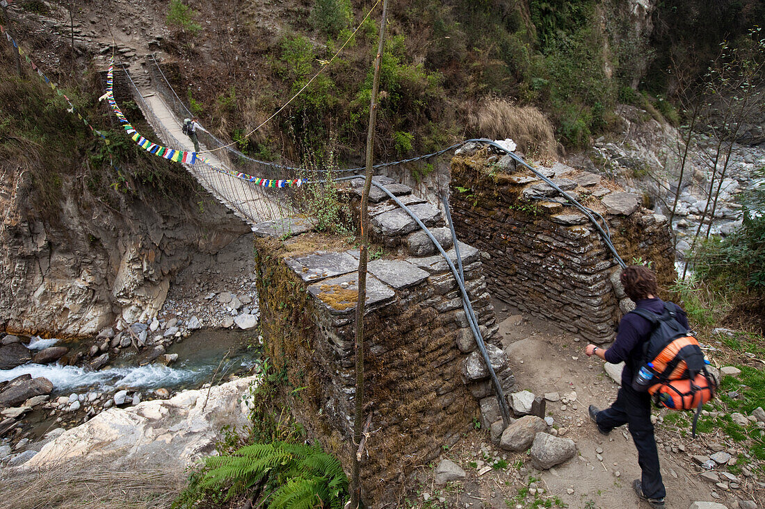 A male trekker approaches a footbridge while a female trekker pauses on the bridge ahead Annapurna Conservation Area, Nepal