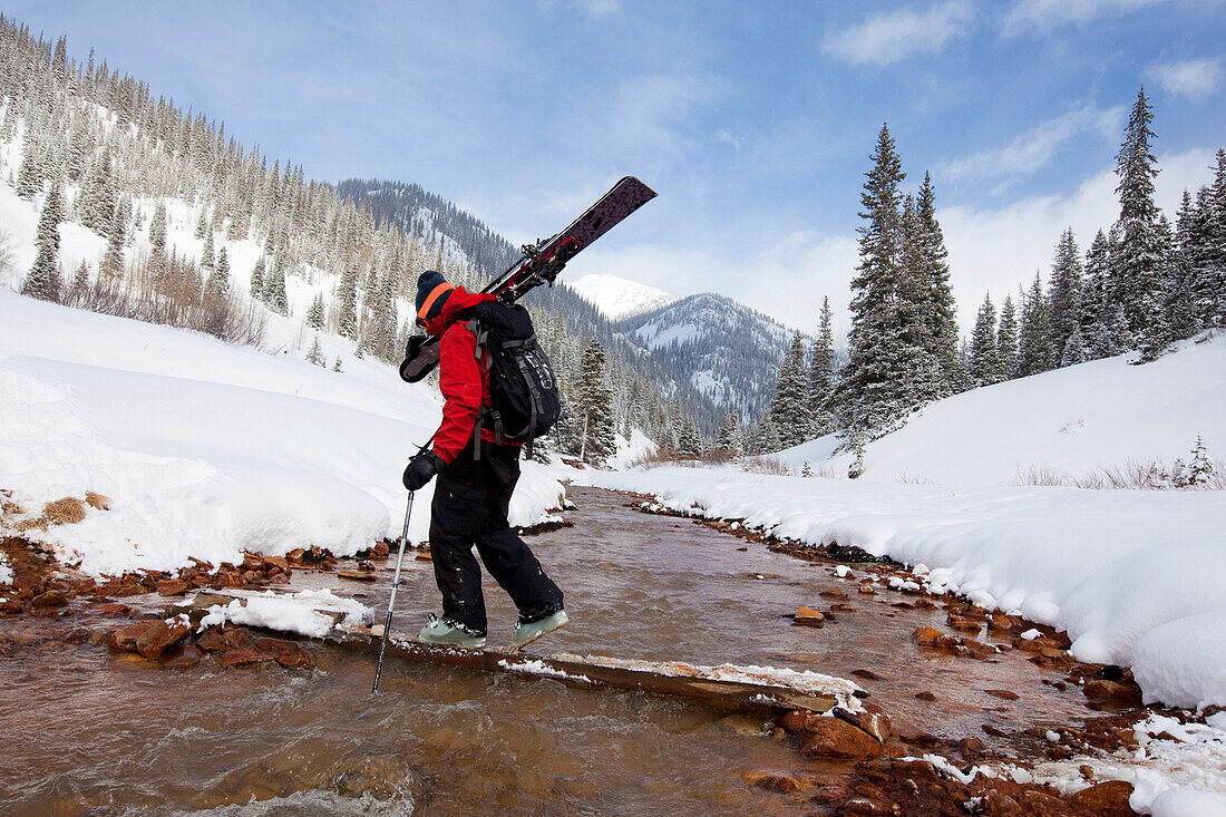 A  man carries his skis across a snowy, mountain stream Colorado, USA