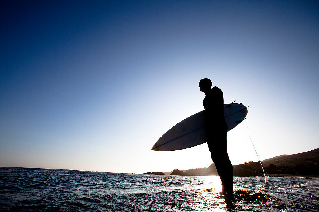A male surfer stands on a rock while being silhouetted by the setting sun Malibu, California, United States of America