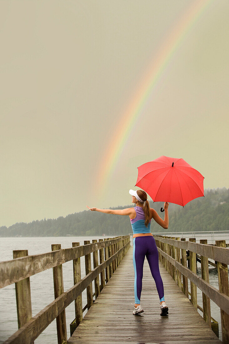 Caucasian woman in sportswear with red umbrella on pier catching rainbow, Bainbridge Island, Wa, USA