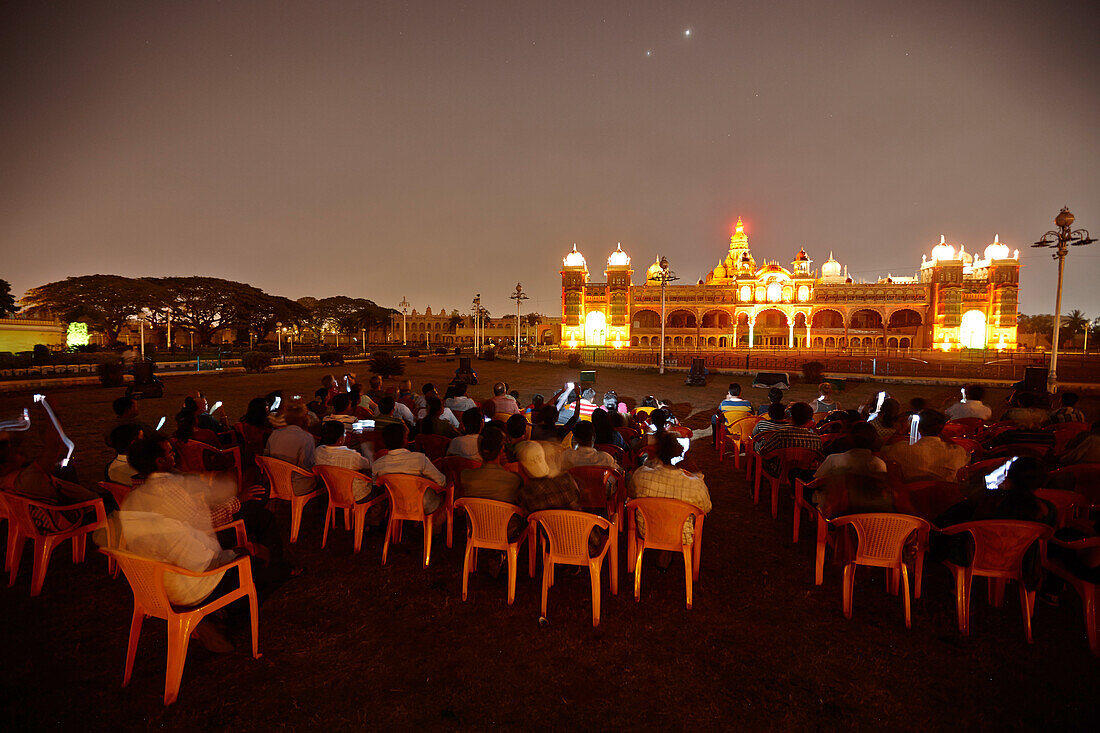 Besucher mit Fotohandys betrachten beleuchteten Amba Vilas Palast am Abend, Mysore, Karnataka, Indien