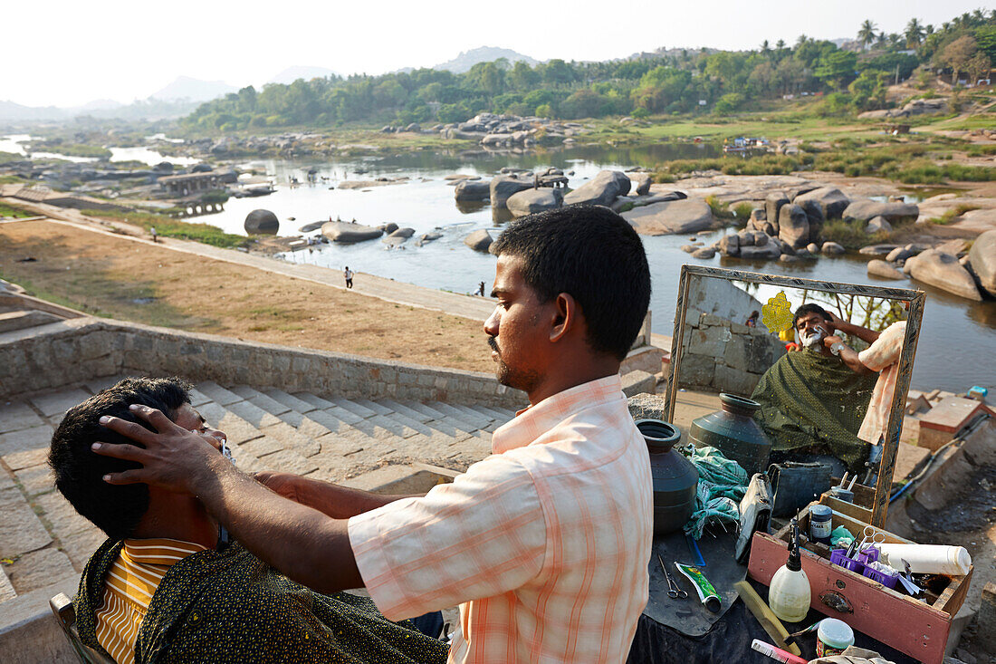 Frisör rasiert Kunden mit Blick auf Gaths am Tungabhadra Fluss, Hampi, Karnataka, Indien