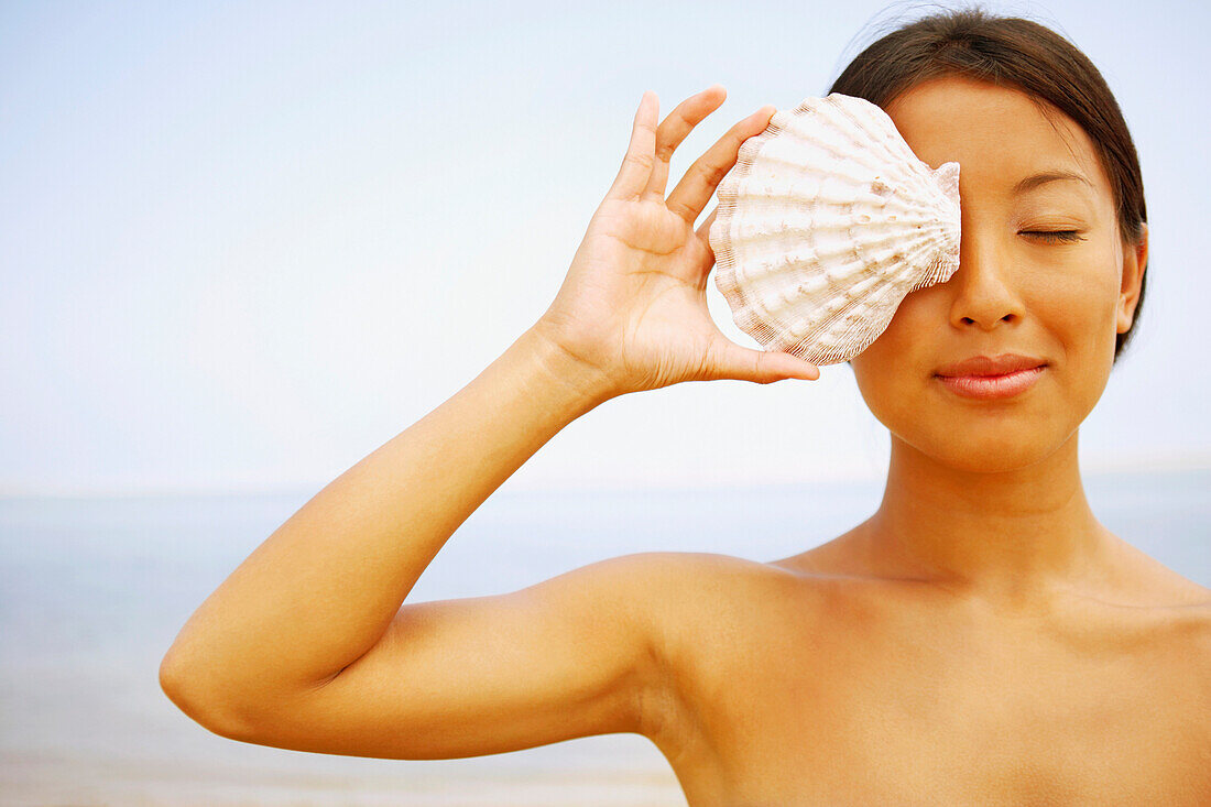 Asian woman holding seashell over eye, Cape Cod, MA