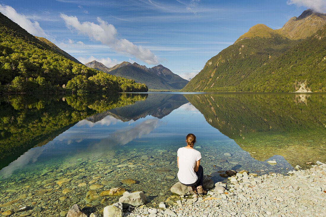 Reflection of the mountains in Lake Gunn, young woman sitting at the lake shore, Southland, South Island, New Zealand