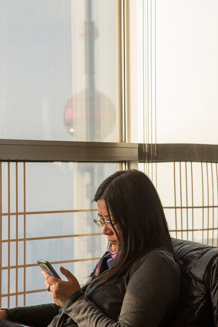 Frau mit Smartphone im Grand Café des Grand Hyatt Hotel im 54. Stockwerk des Jin Mao Tower Hochhaus, Pudong, Shanghai, China