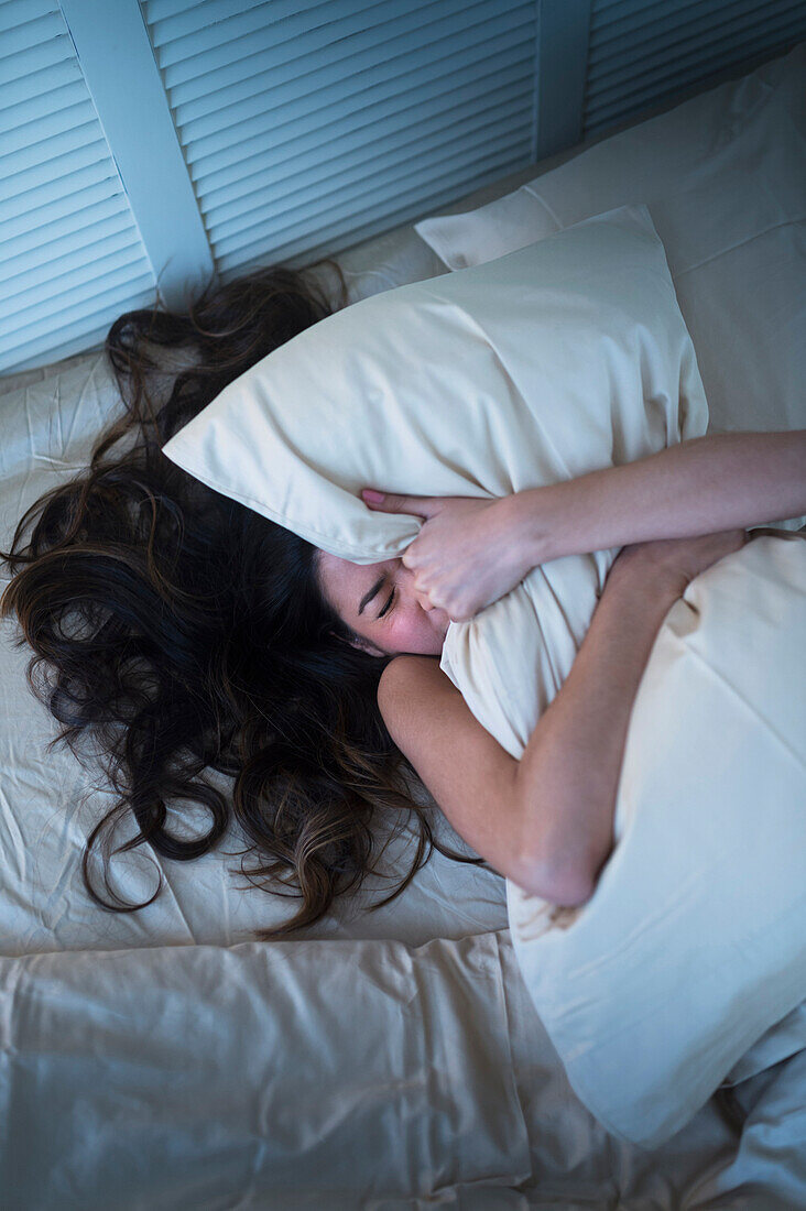 Frustrated Hispanic woman gripping pillow in bed, Jersey City, NJ, USA