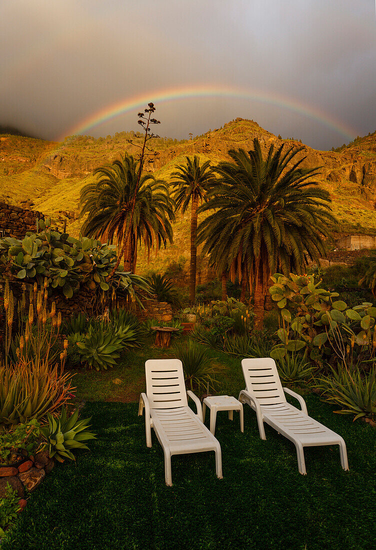Deck chairs in a garden, rainbow behind palm trees, Valley of El Risco near Agaete, Natural Preserve, Parque Natural de Tamadaba, UNESCO Biosphere Reserve, West coast, Gran Canaria, Canary Islands, Spain, Europe