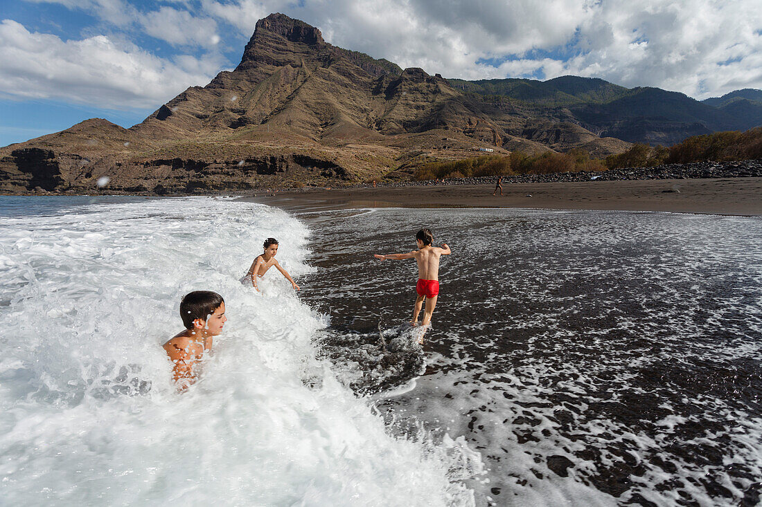 Kinder spielen in den Brandungswellen, Berg Faneque und Playa del Risco, Strand, bei Agaete, Atlantik, Naturschutzgebiet, Naturpark Tamadaba, UNESCO Biosphärenreservat, Westküste, Gran Canaria, Kanarische Inseln, Spanien, Europa