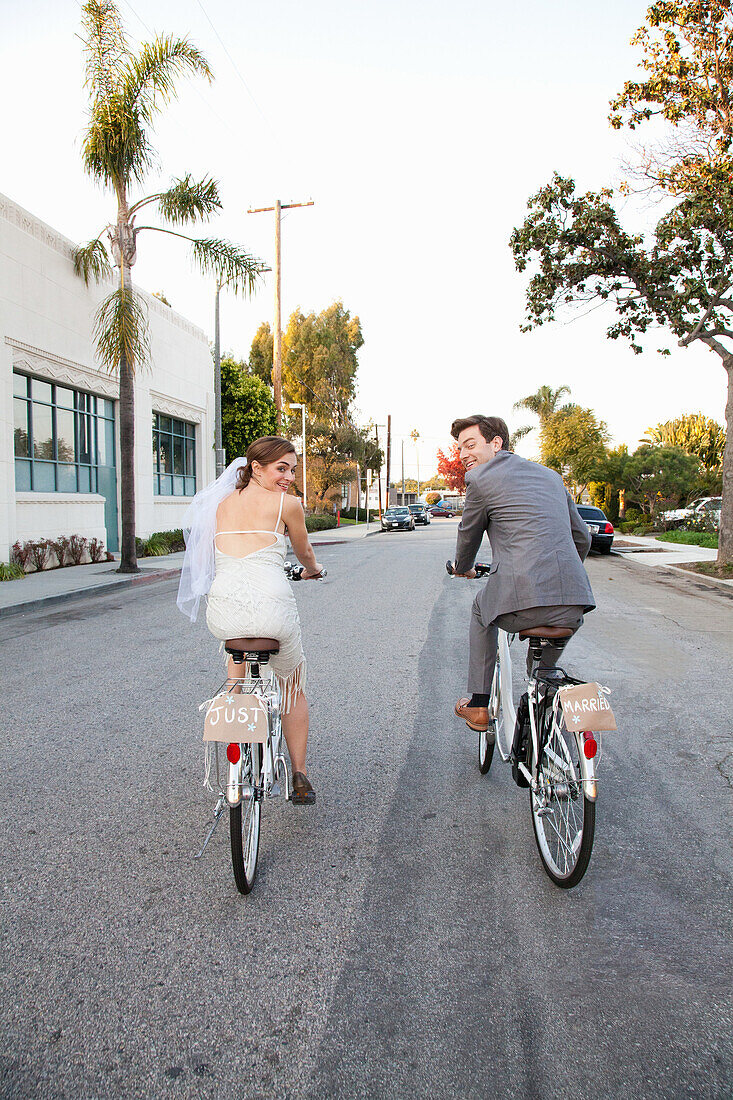 Young newlywed couple cycling along street, rear view