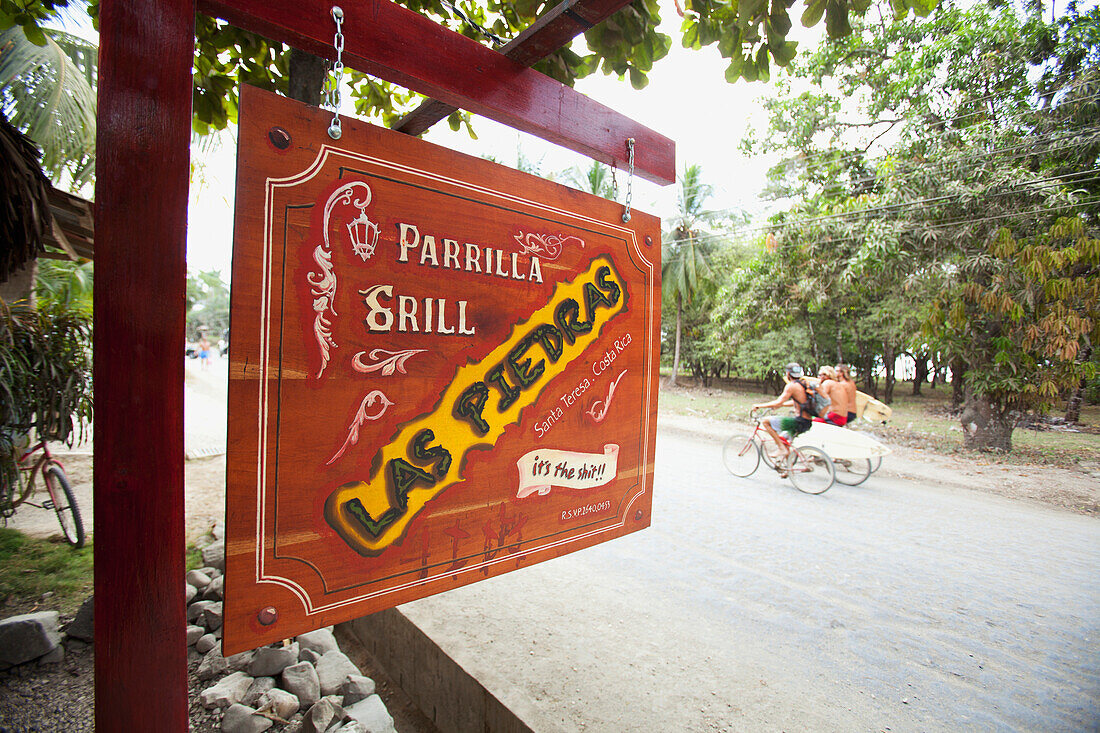 'The Parrilla Grill, Las Piedras Restaurant Sign Hanging And Surfers Ride Past On Their Bicycles In Santa Teresa And Mal Pais (Malpais) On The Nicoya Peninsula; Puntarenas Province, Costa Rica'
