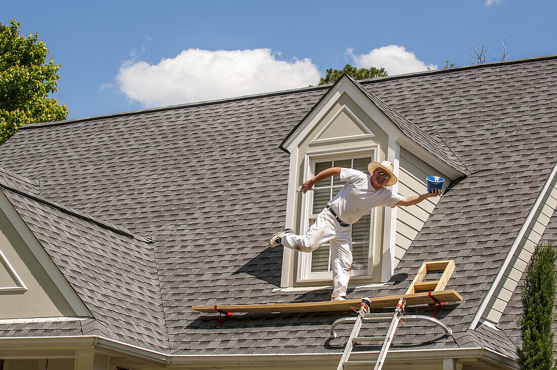 A 45 year old man on a roof balancing on one leg clowning around while painting a house.