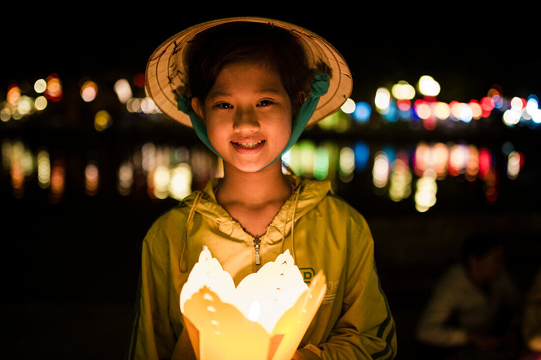 Vietnamese young girl along the river with typical vietnamese conic hat holding a typical lantern with other blurred lanterns light in the background, Hoi An, Vietnam
