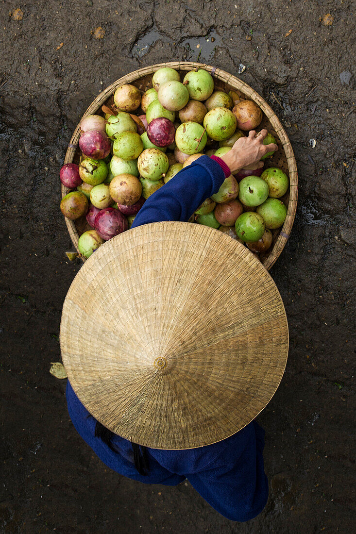 Woman with typical vietnamese conic hat moves vegetables on her circular basket on the floor along a street market in Vietnam