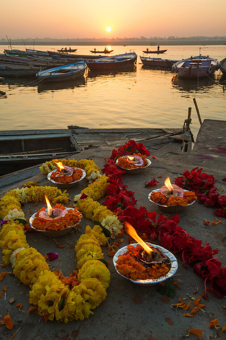 Lights and garlands of flowers on the banks of Ganges river at sunrise with boats on the river and rising sun in the background, Varanasi, India