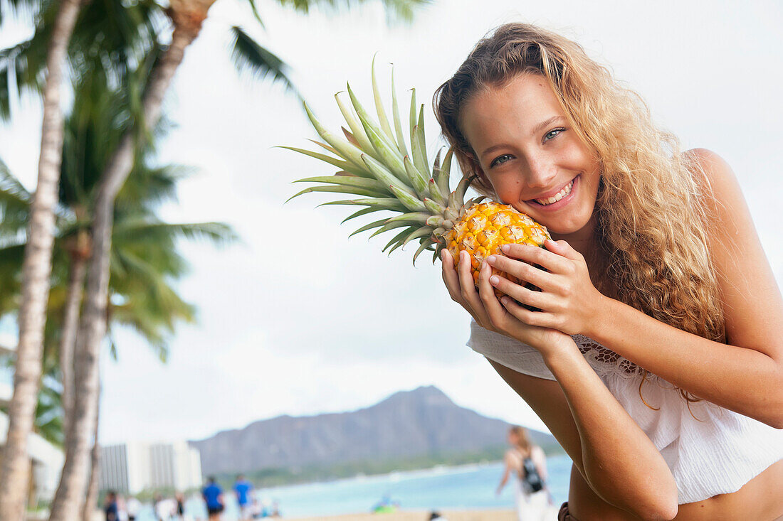'A young woman poses with a fresh pineapple and Diamond Head in the background; Oahu, Hawaii, United States of America'