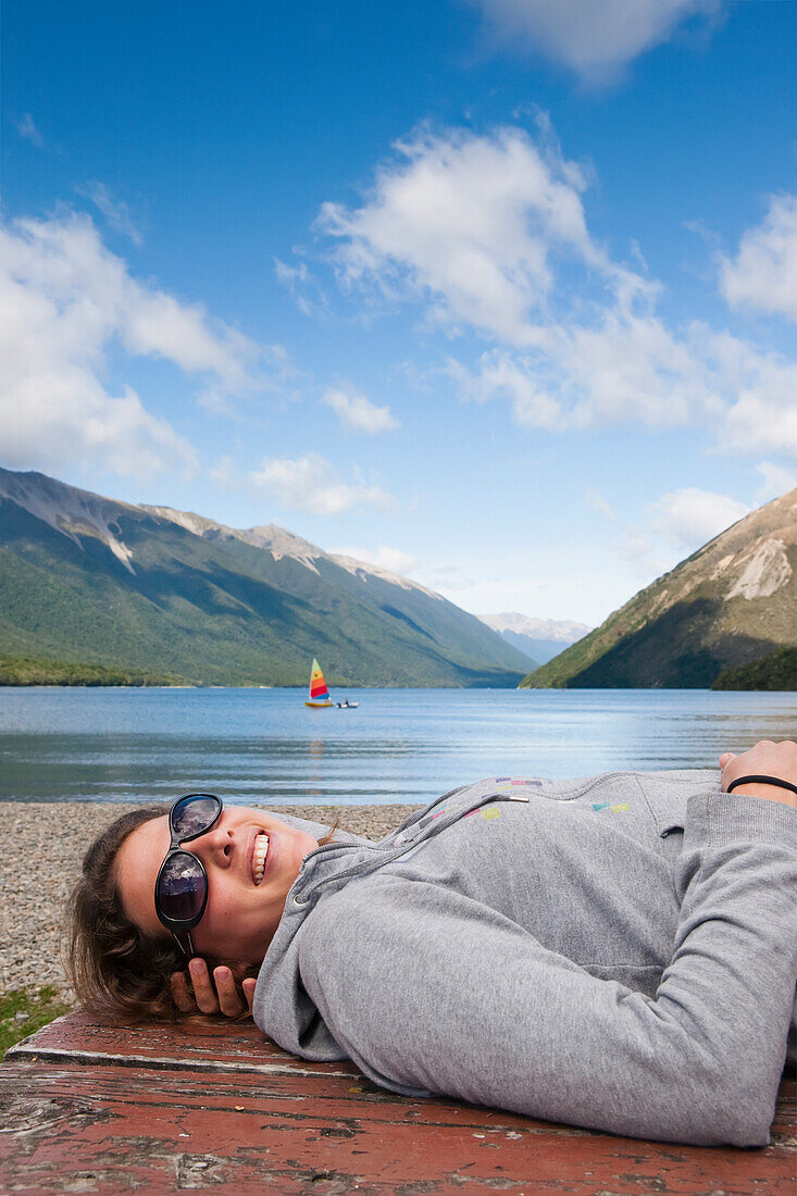 'A Girl Lies Down On A Picnic Table And Enjoys The Sun With A Sailboat In Lake Rotoiti In The Background, Tasman Region; New Zealand'